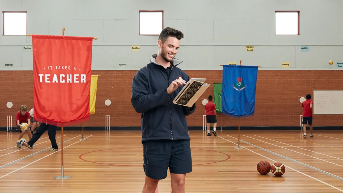 A gym teacher carries a Chromebook with its screen flipped open into tablet mode as he walks across a gym with children playing in the background.