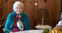 Photograph of an old lady, with white hair, dressed in green and purple, sitting in front of a table and smiling at the camera.