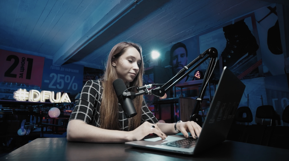 A woman at a laptop, sitting in a studio next to a large microphone.