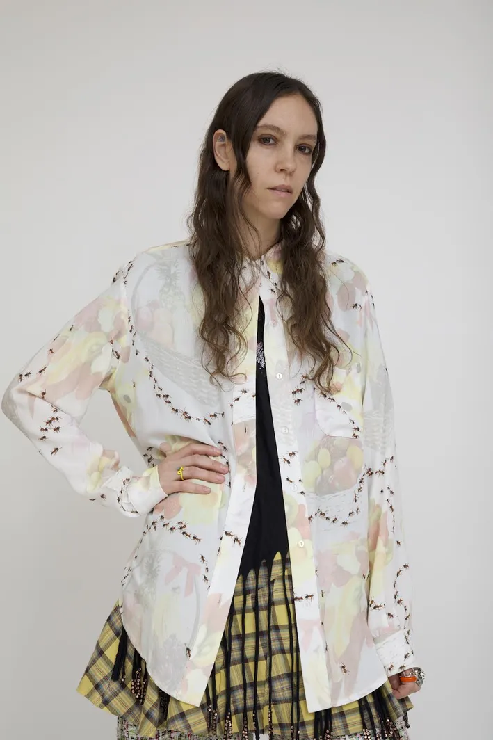 Headshot of female designer Hillary Taymour posing against a white background with a buttoned down shirt and yellow striped skirt