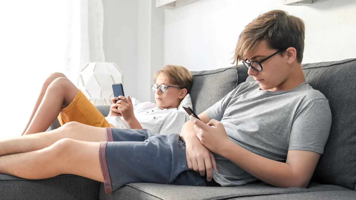 Two boys, one child and one teen, sit on a couch looking at their mobile phones.