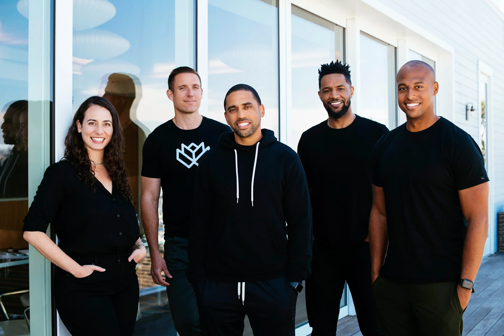 Five team members stand near a glass door and smile in the camera. They are all wearing black tops and black pants.