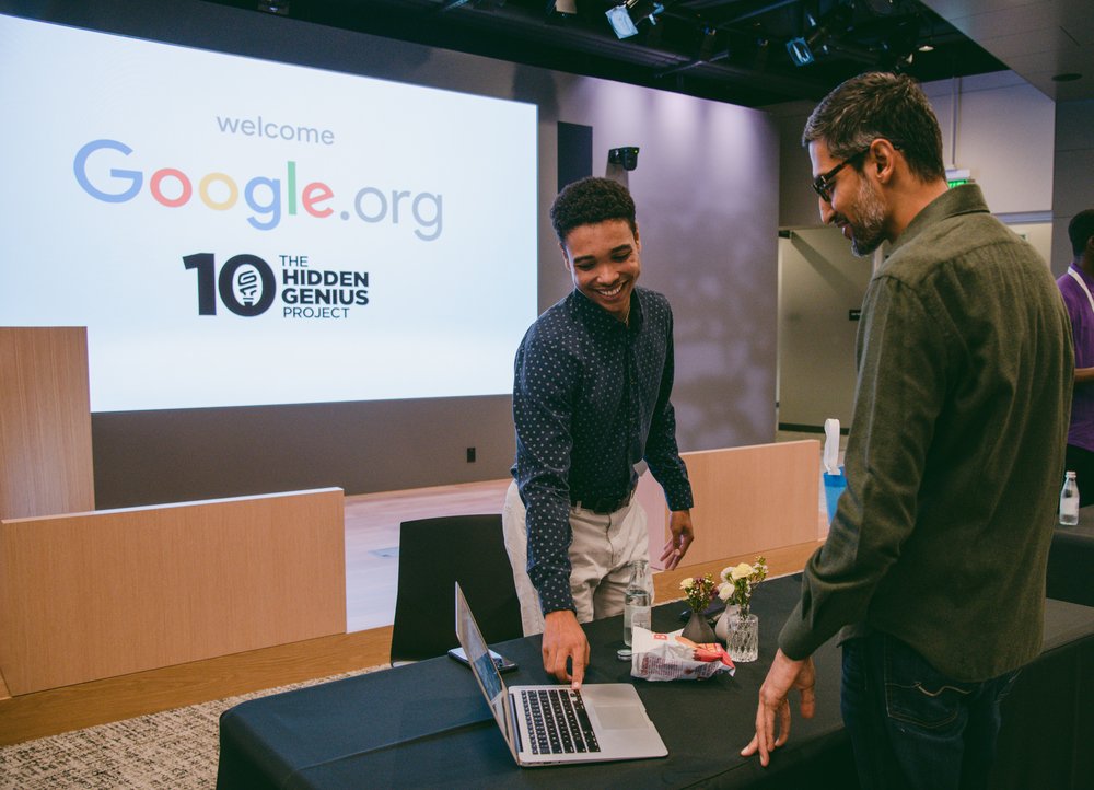 computer science - Student Ian stands behind a desk operating a laptop to demonstrate his platform for Sundar, who is standing to the right of the desk. Both are smiling. A screen in the background shows the Google.org and The Hidden Genius Project logos.