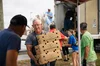 Eight volunteers are pictured taking food from a truck and carrying it towards the camera. A man with white hair and glasses is carrying three cardboard boxes in the foreground, while other volunteers pass boxes along a line in the background.