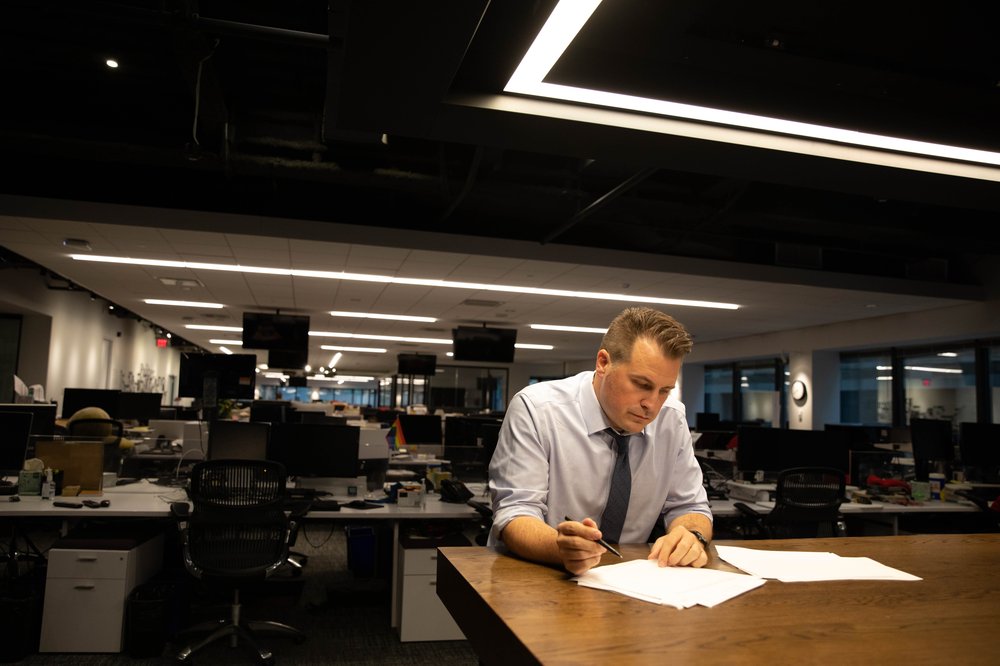 Brendan wears a light shirt and dark tie and sits at a long wooden table, editing a news article with a pen.