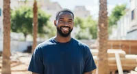 A photo of a black man with short hair and beard smiling. In background there are blurry trees and buildings.