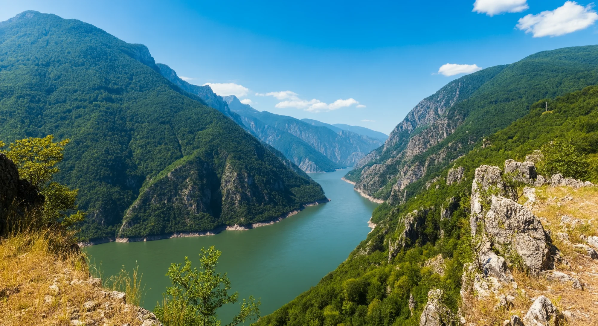 Une large rivière serpente à travers une gorge profonde creusée dans une chaîne de montagnes verdoyante et luxuriante sous un ciel bleu clair. La rivière est calme et reflète le paysage environnant. Le soleil brille de mille feux, projetant des ombres sur les pentes et mettant en valeur les textures des rochers.