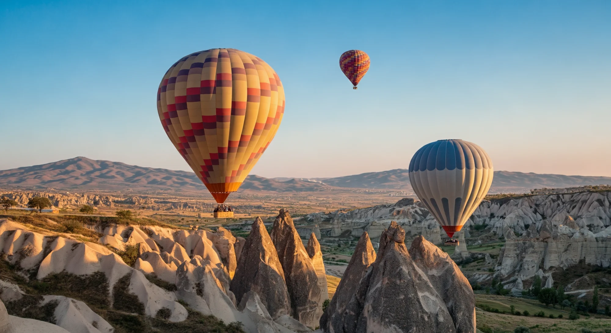 Trois montgolfières flottent dans le ciel au-dessus d’un paysage accidenté de formations rocheuses. Les ballons sont colorés et ont un panier suspendu en dessous. Le soleil brille et le ciel est bleu.