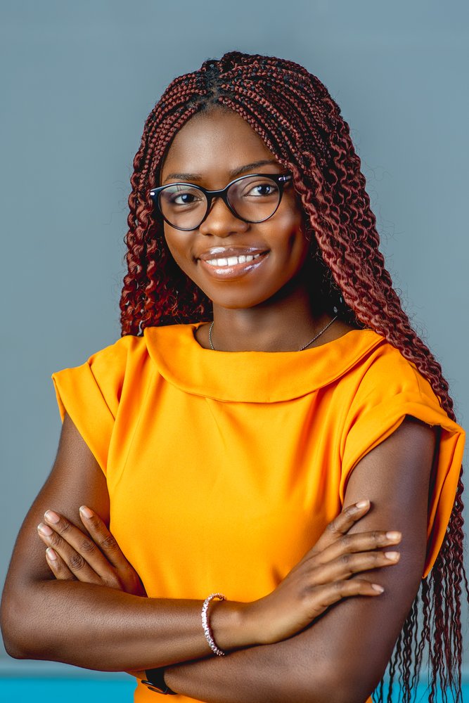 Oiza, with her arms crossed and wearing black glasses and an orange top, smiles at the camera for a headshot image.