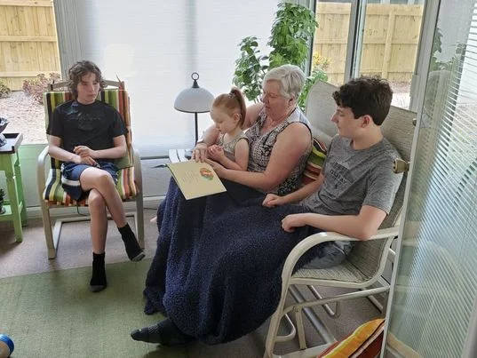 Image of an elderly woman reading a book while holding a toddler on her lap and with two teenagers seated next to her.