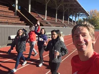 A selfie of six people walking on a red, outdoor track. They are smiling at the camera, waving and holding up peace signs.