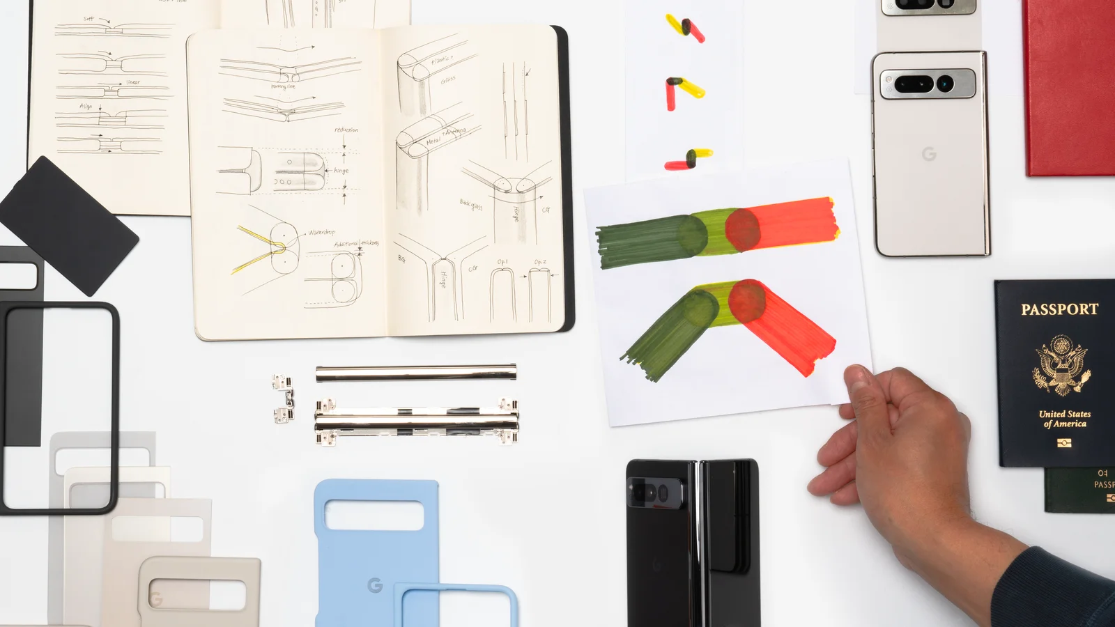 An overhead shot of various items laying on a white surface, including a passport book, a Pixel Fold, casings for the Pixel Fold in various colors, mechanical parts, two open sketch books featuring drawers of hinges. A hand is in the shot, laying down a pl