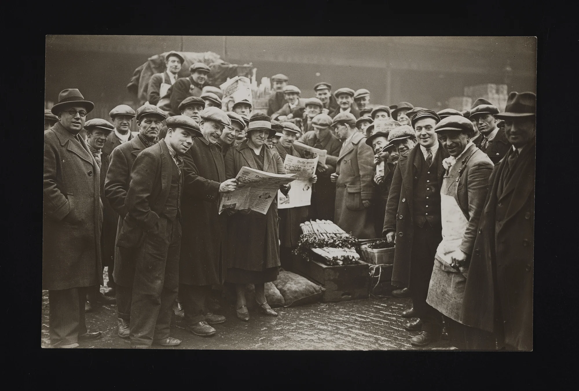 A black and white photograph of a group of people standing reading the Daily Herald