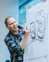 Image shows a person in a plaid shirt holding a microphone, leaning around and looking at an easel with a canvas on it that shows a drawing of two cartoon characters. The person is talking into the microphone.