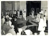 Black and white photo of a large crowd of people in a room watching two men standing in the center with a microphone stand.