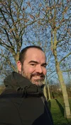 photo of a man with a beard wearing a black jacket, smiling in front of trees and a clear sky