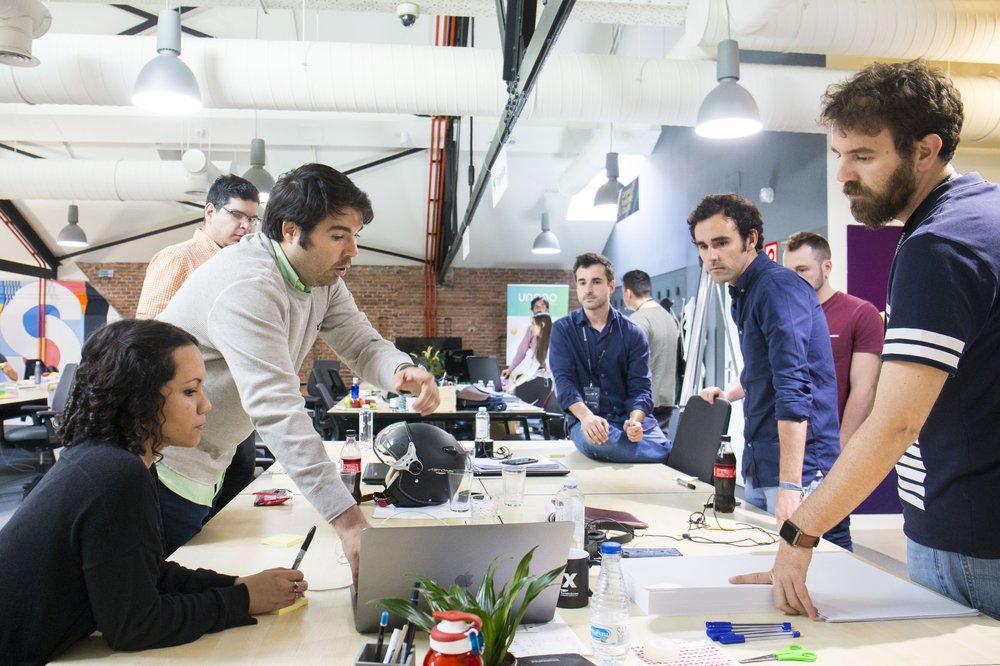 A group of people standing around a laptop computer, which is sitting on a desk. One man in a gray sweater is speaking and the rest of the group is listening.