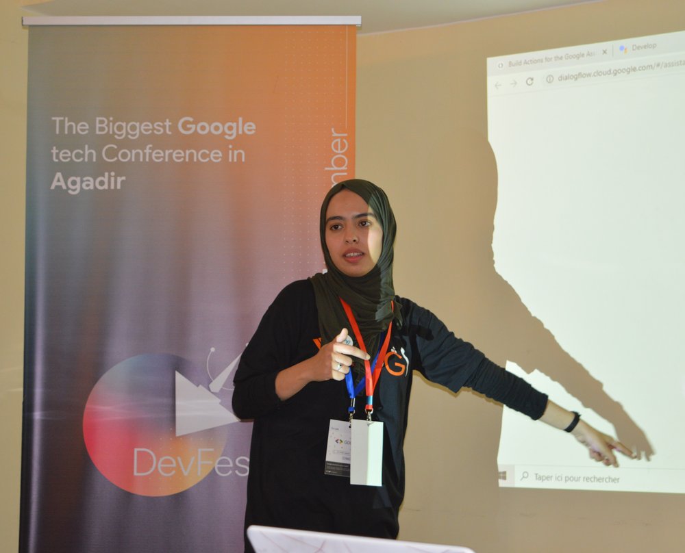 A photo of Hanane, wearing a black GDG t-shirt and gray head cover, giving a talk at GDG Agadir DevFest. To her left is a banner that reads, “The Biggest Google tech Conference in Agadir: DevFest.”