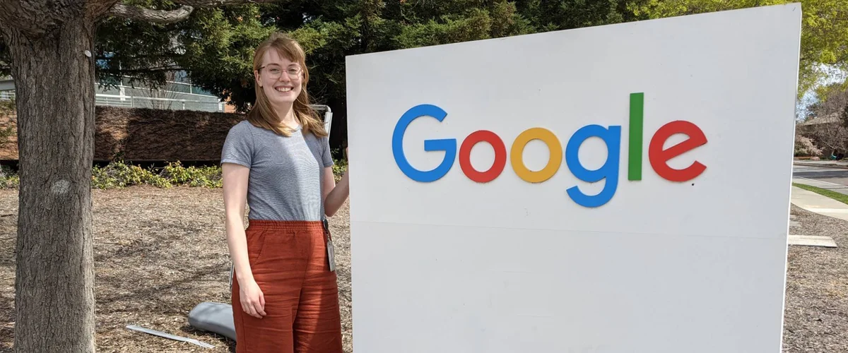 Woman standing next to a Google sign. On her left is a tree trunk, and behind her are trees, a building and a low brick wall.