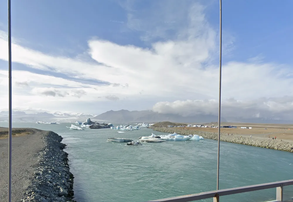 Shown is a view from a bridge overlooking a river, where icebergs float in the water. The river is flanked by rocky banks.