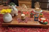 A wooden platter on a table with a red tablecloth. On the platter include various lamps and flowers