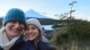 Selfie de dos mujeres en abrigos pesados y gorros de lana, frente a una montaña cubierta de nieve al atardecer.