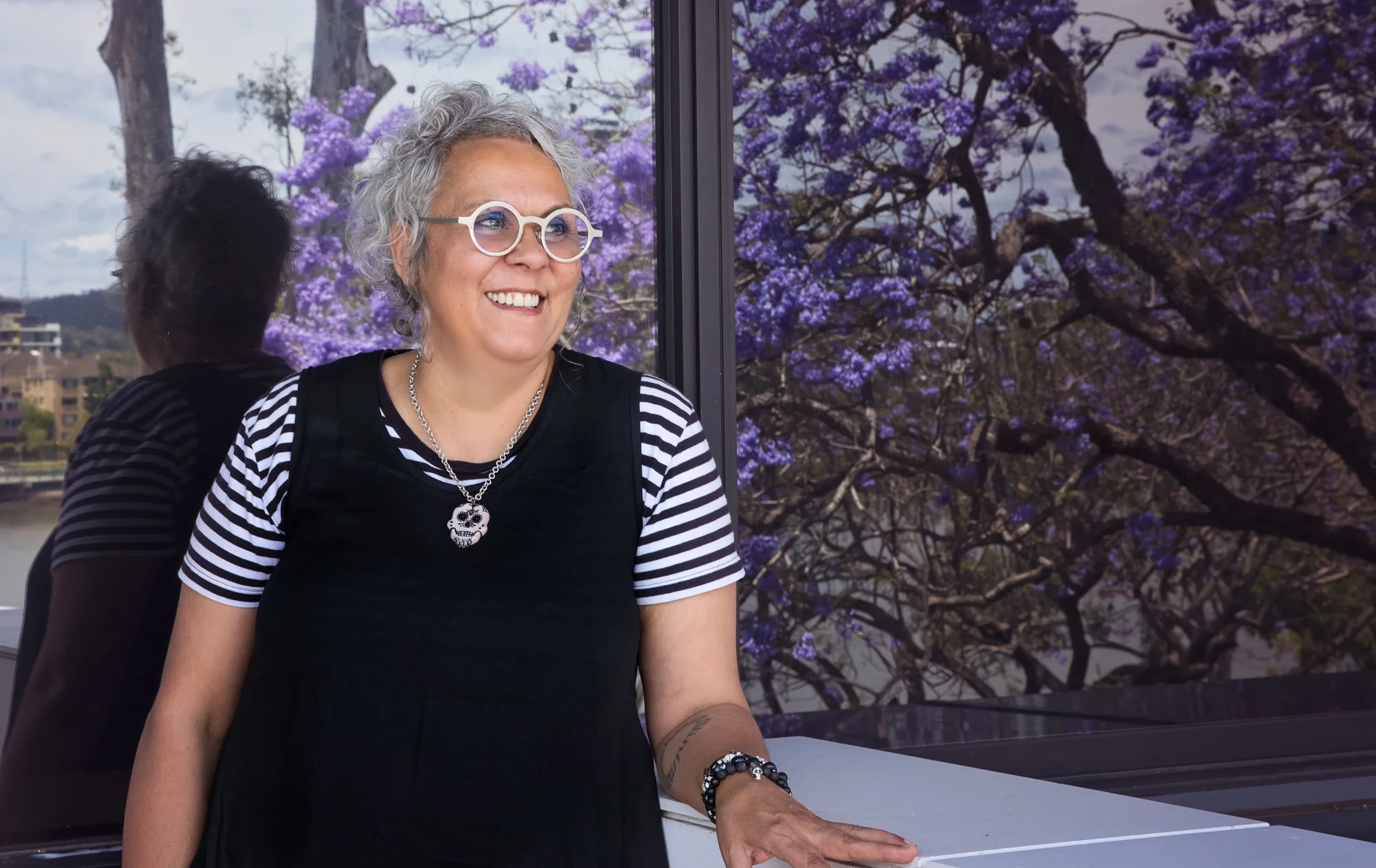 A photograph of a woman with gray hair and round white glasses smiling and looking to the right, with purple flowers behind her.