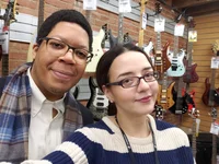 A man with glasses looks into the camera smiling. A woman with glasses next to him is also looking into the camera and smiling, and she is holding the camera taking a selfie. They are in a guitar store.