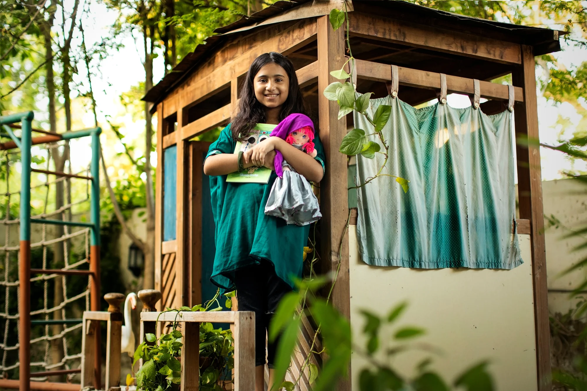 A photograph of a girl in a teal shirt holding a book and a doll, with trees and a wooden structure behind her.