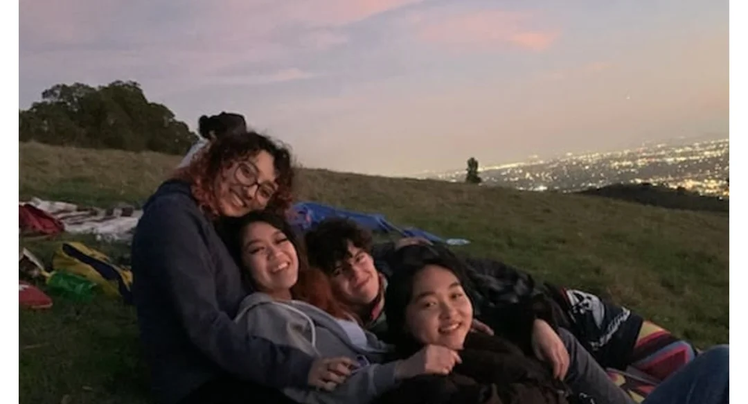 Emily Ronquillo-Ruiz and three other students huddle on a park blanket with a city view at twilight behind them