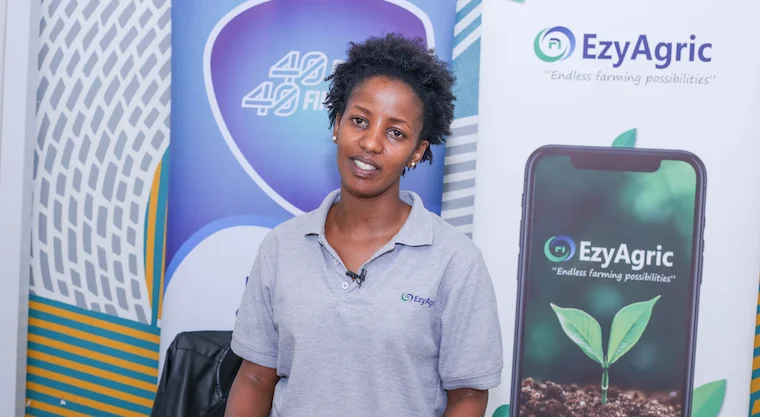 A woman in a light blue polo shirt stands in front of a sign that reads "EzyAgric."