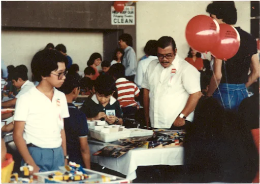 Paolo sitting at a table building a Lego set in a room full of others also focused on their Lego creations. To his right, a man stands over his shoulder watching him build the set