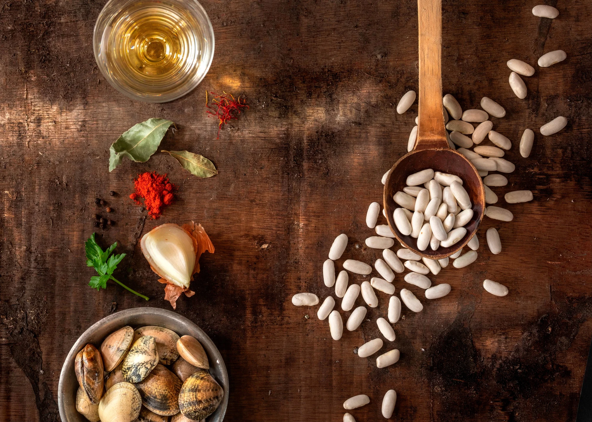 Ingredients are placed on a wooden table: on the right, beans on a wooden spoon; on the left from top to bottom: a glass with oil, saffron threads, bay leaf, paprika, peppercorns, a clove of garlic, parsley and clams.