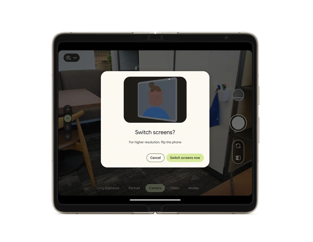 A screenshot of the Fold’s display showing camera mode opened and the lens is looking at an office space. A box on the screen shows an illustration of a person and a frame and underneath this illustration it reads “Switch screen? For high resolution, flip the phone.” Underneath this are two option boxes that read: Cancel and Switch screens now.