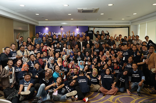 A large group poses for a photo with a banner that reads “Trusted Media Summit 2018” in the background