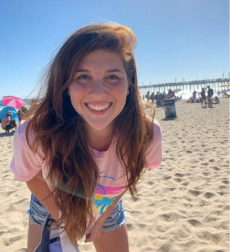 Image showing a young woman looking into the camera smiling. She has long brown hair and she's wearing a pink shirt and shorts. She's standing on a beach, in the sand, there is blue sky behind her.