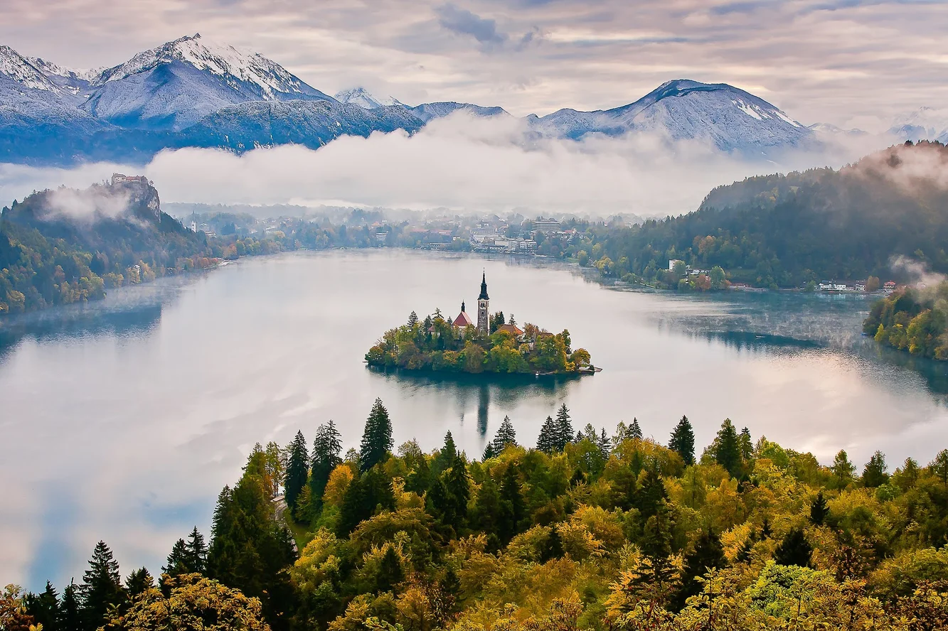 A picture of a large lake surrounded by trees and against a backdrop of ice-capped mountains. In the middle of the lake is a small island with a few buildings on it surrounded by trees.