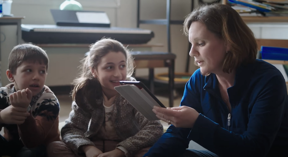 A mother looking at a tablet and reading aloud to two children.