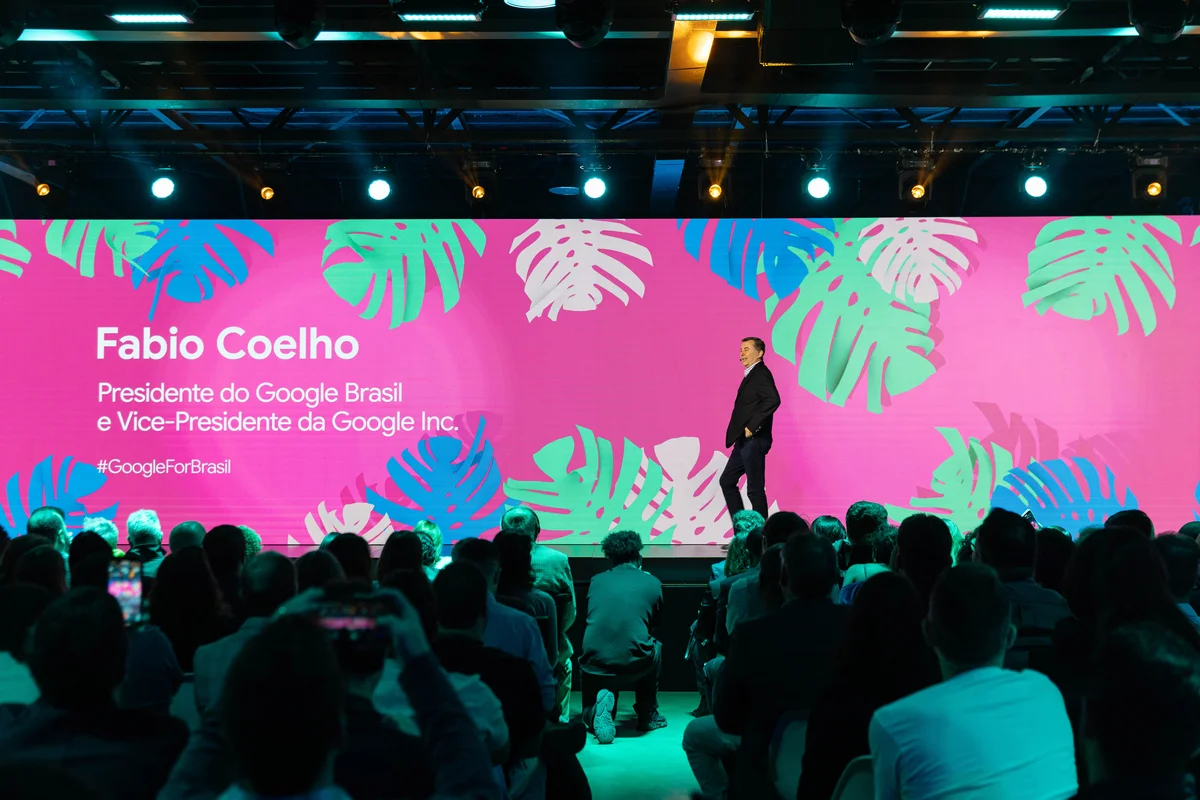 Fabio Coelho, um homem branco de terno escuro, está num palco em frente a uma audiência. No fundo, uma tela de fundo de rosa e plantas com o nome e título do presidente do Google Brasil