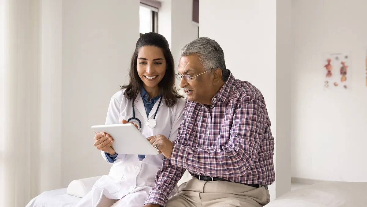 A doctor sits next to a patient in a medical appointment room. The doctor is holding a tablet in her hand. The patient is touching the tablet with his left hand.