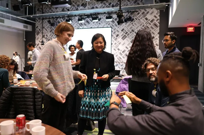 The image shows people engaged in conversation at a research workshop in a modern room with a patterned wall and a large screen. In the foreground, a woman in a multicolored sweater and another in a patterned dress with a black blazer are smiling and hold