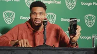 NBA player Giannis Antetokounmpo behind a microphone wearing a burnt orange hoodie and holding a black Pixel phone with glass of water on his right in front of green backdrop with text ‘Google Pixel’