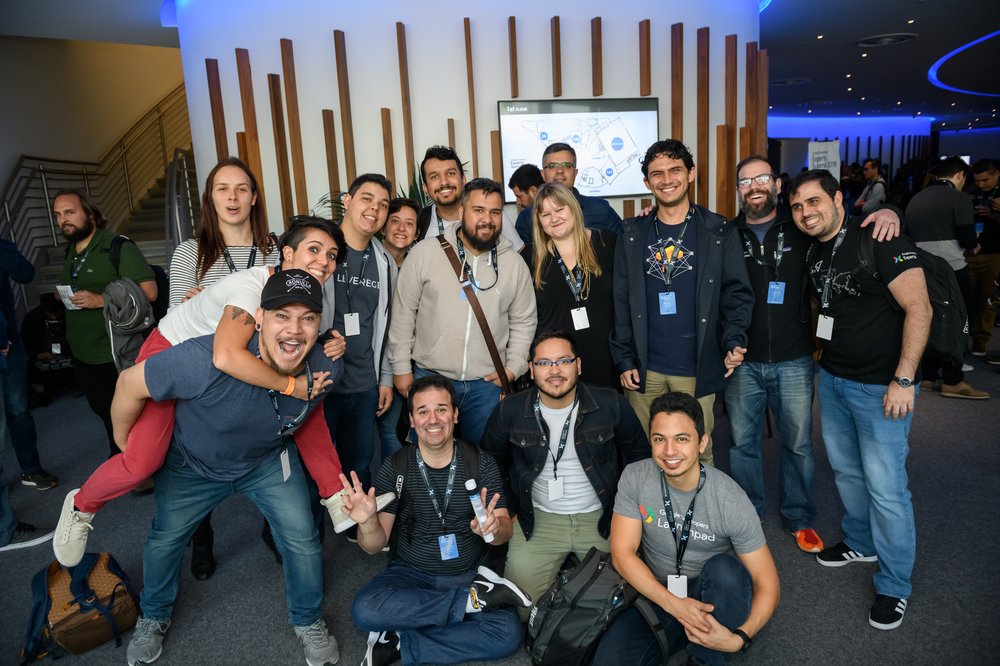 A group photo of Google developer community members at an event, wearing lanyards and smiling at the camera. Some have their arms around each other, and one is giving another a piggy-back ride.