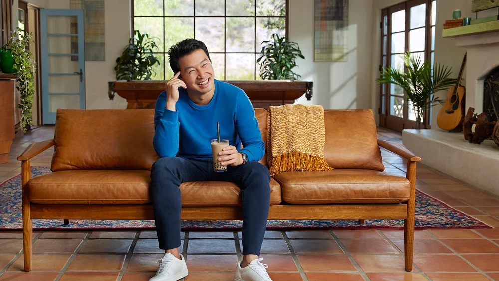 Simu Liu sitting on a brown couch in a living room setting