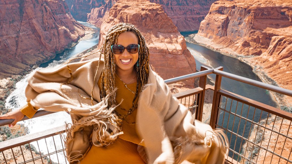 A woman in long braids wearing a tan scarf and sunglasses poses on a viewing stand over a dramatic natural canyon.
