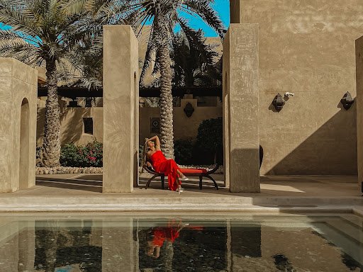 A woman in a red dress reclines on a chaise in front of a pool and palm trees in a courtyard.