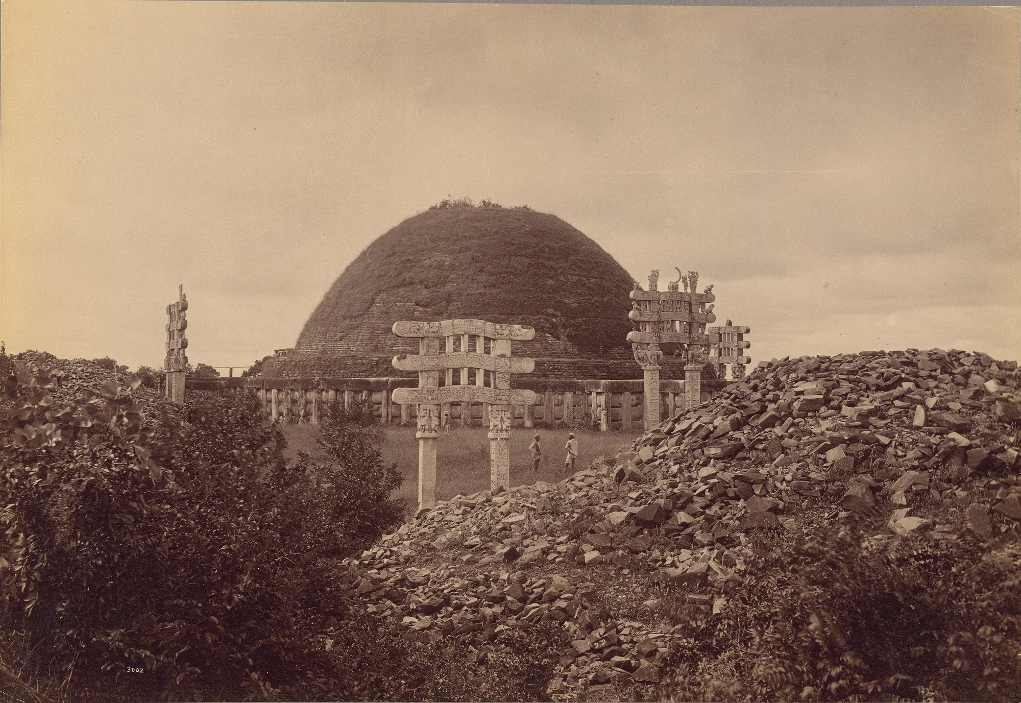 Black and white photograph of a large outdoor mound of earth, with tall stone edifices in front of it.
