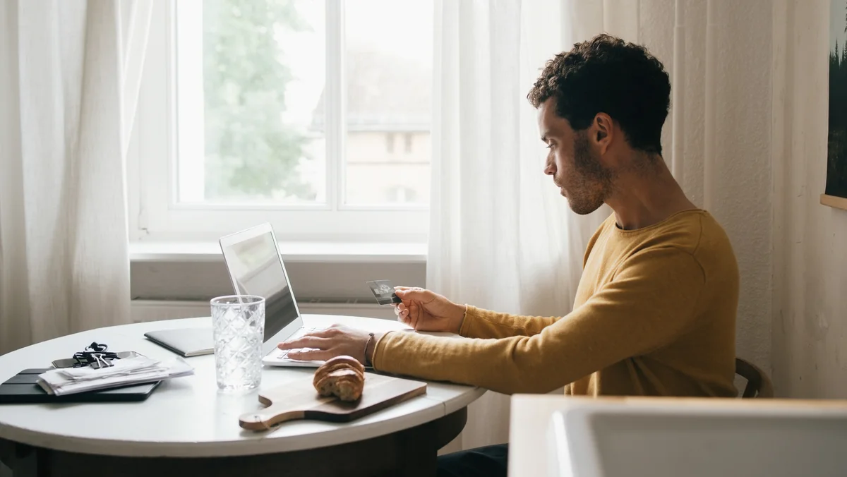 Youtube Advertising - A young man wearing a yellow, long-sleeved shirt sitting at his kitchen table holding a credit card and typing on a laptop computer.