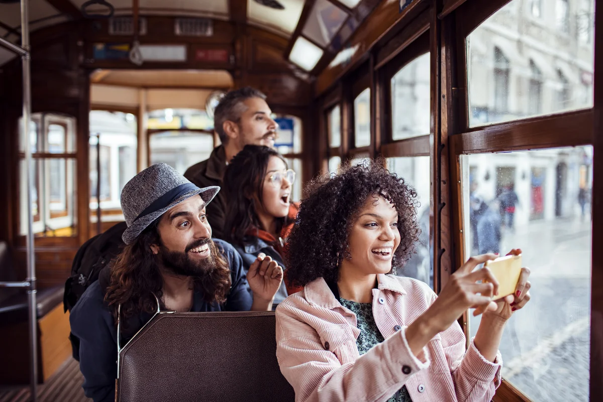 A group of people sitting in a tour bus. All are looking out the window with excited expressions, and the woman in the front is taking a picture with her phone.