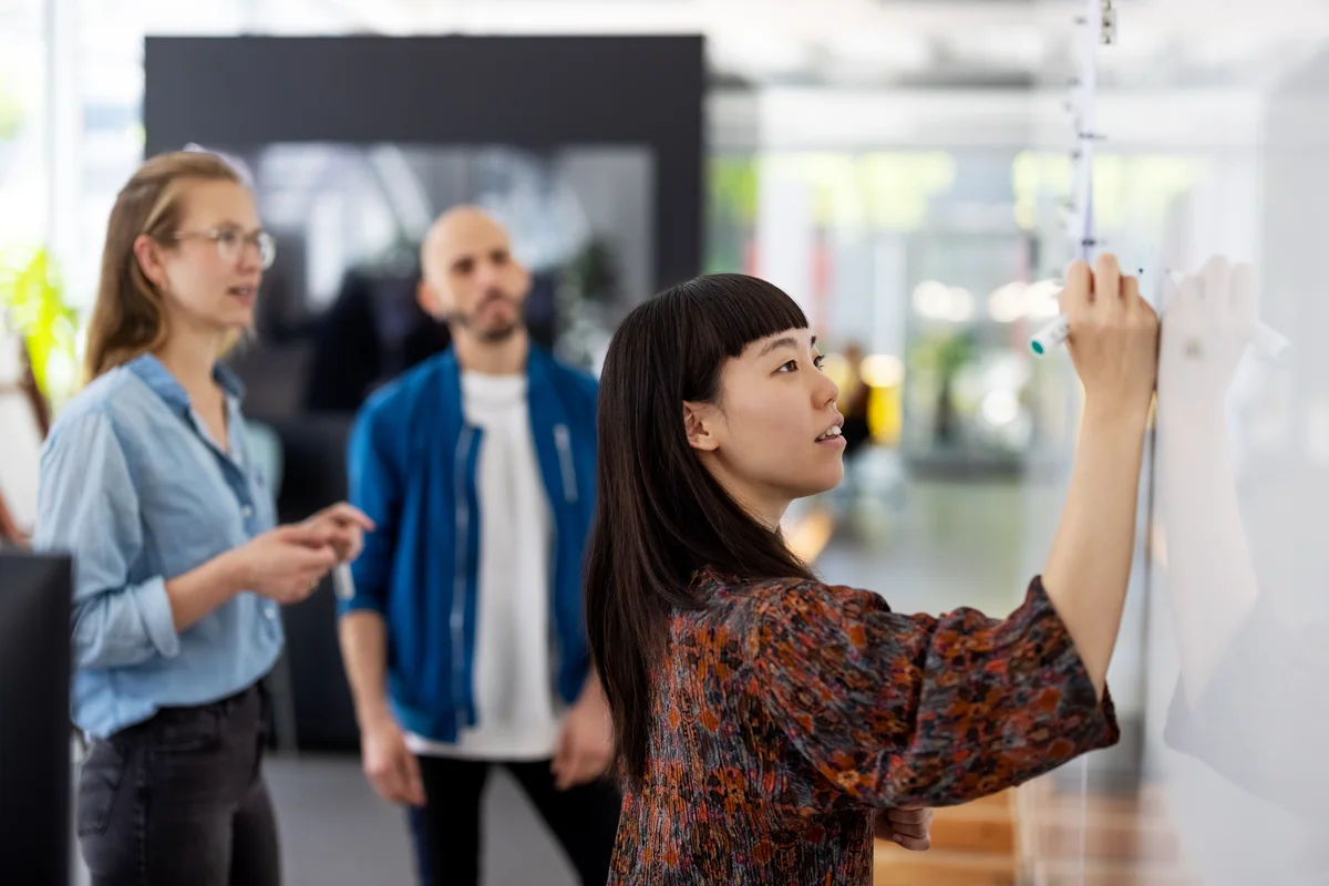 Two women and one man looking at a whiteboard. One of the women is writing on the whiteboard.